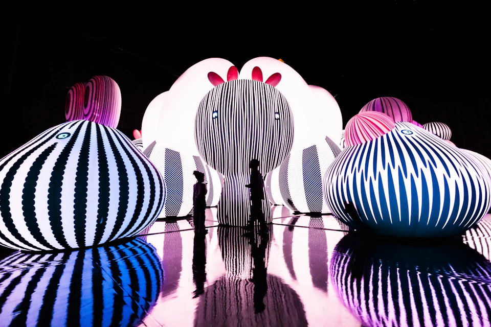 Silhouettes of two people walking among large, illuminated, striped inflatable sculptures with a reflective floor.