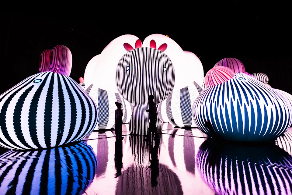 Silhouettes of two people walking among large, illuminated, striped inflatable sculptures with a reflective floor.