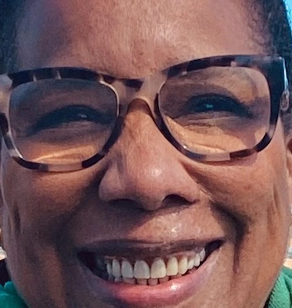 A person wearing glasses, a green scarf, and a dark coat stands on a balcony with a cityscape and blue sky behind them.