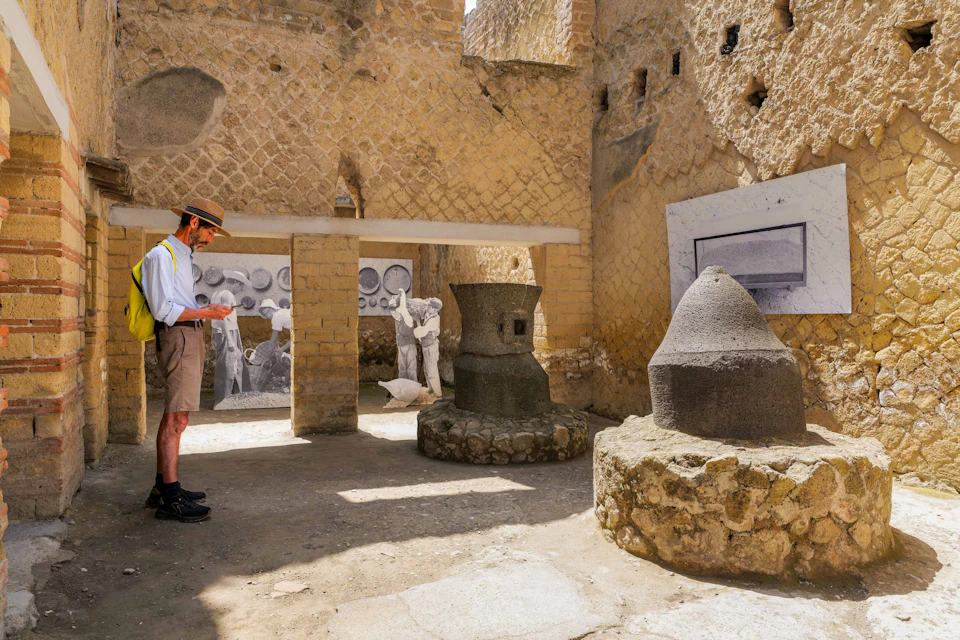 A person wearing a hat and shorts examines ancient ruins with stone artifacts and historical murals on the walls.