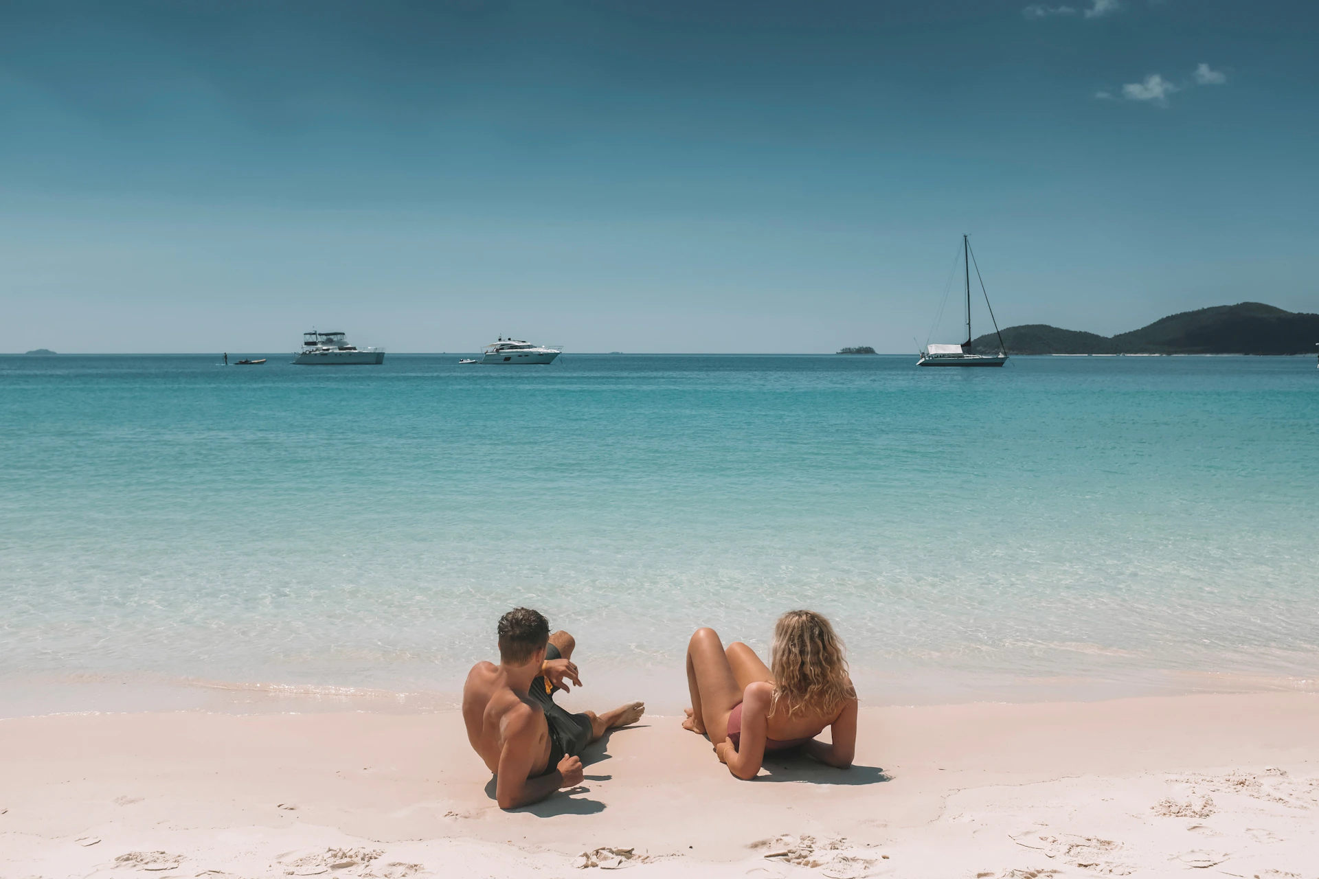 A couple and a child sit on a sandy beach facing a calm blue sea with several boats anchored in the distance.