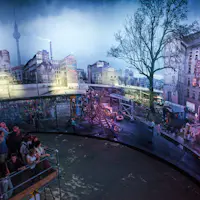 A group of people observe a panoramic display of a cityscape with historical buildings, graffiti, and a large tree.
