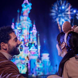 En man och en kvinna ler och tittar på ett starkt upplyst slott och fyrverkerier på natten. Kvinnan bär glittriga musöron.