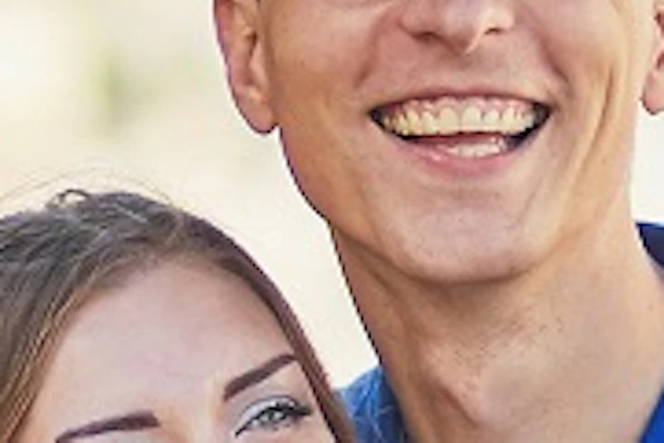 A smiling couple stands on a street, taking a selfie. Trees and buildings are visible in the background, along with a view of the water.
