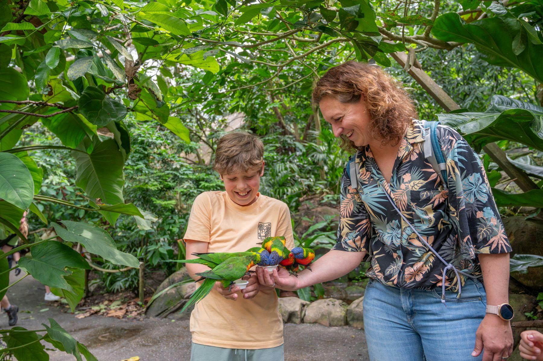 A woman and a boy smile while feeding colorful parrots in a lush, green, outdoor setting.