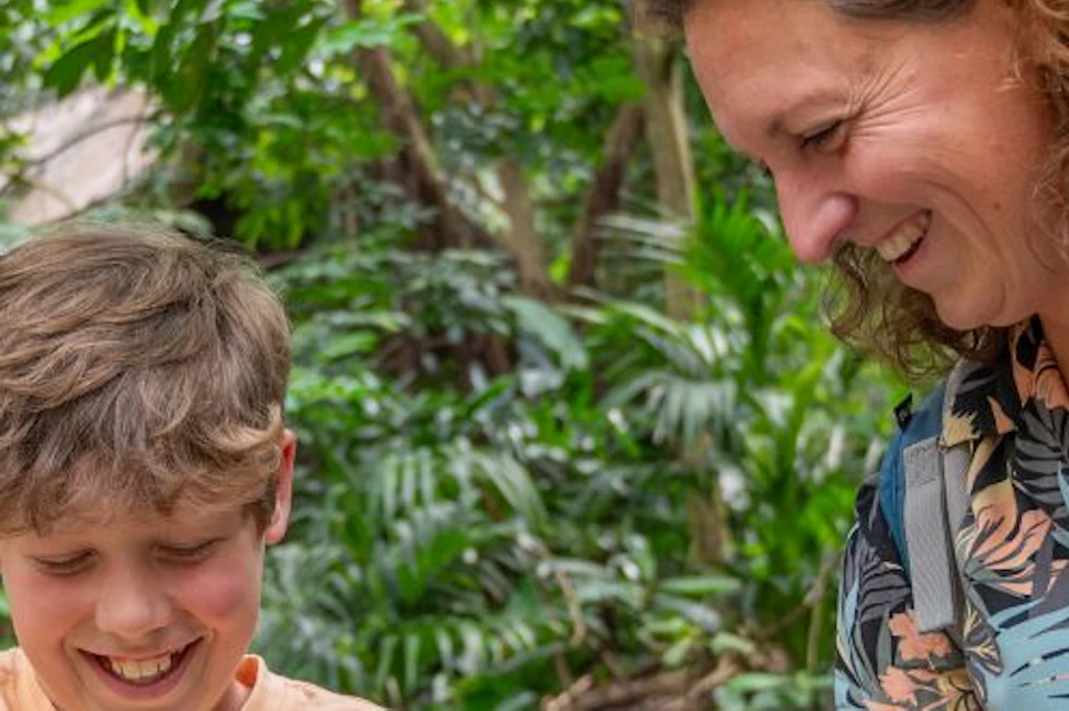 A woman and a boy smile while feeding colorful parrots in a lush, green, outdoor setting.