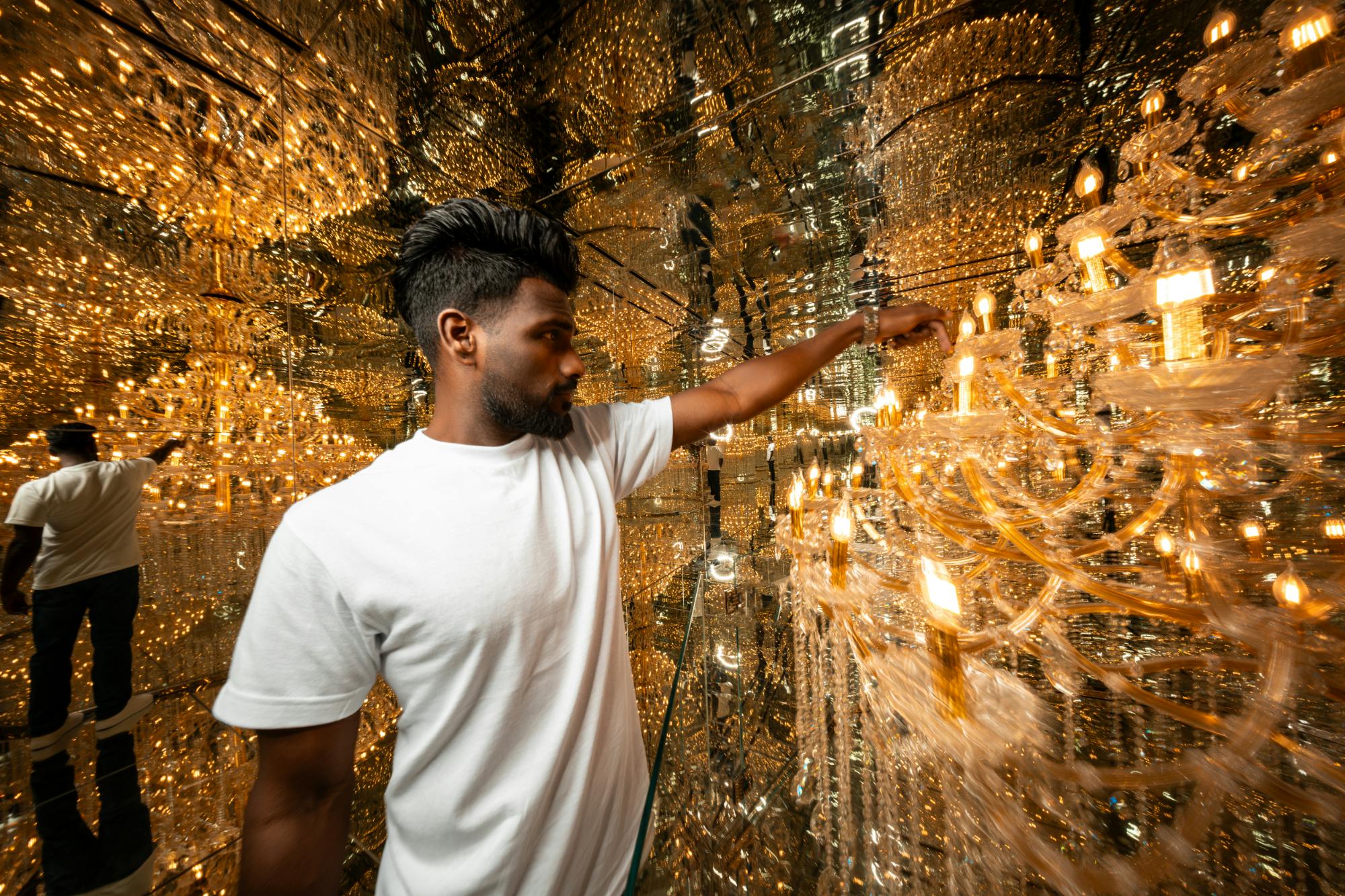 Mężczyzna w białej koszulce stoi w lustrzanym pokoju wypełnionym wiszącymi światłami, tworząc olśniewające, refleksyjne otoczenie.