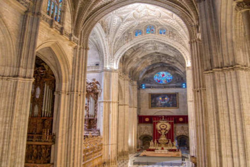 Interior of a cathedral with ornate arches, stained glass windows, a large altar, and intricate architectural details.
