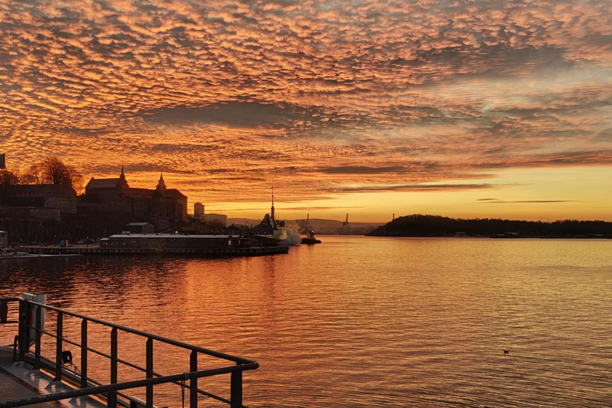 A scenic view of a harbor at sunset with orange clouds, a ship, distant buildings, and a railing in the foreground.