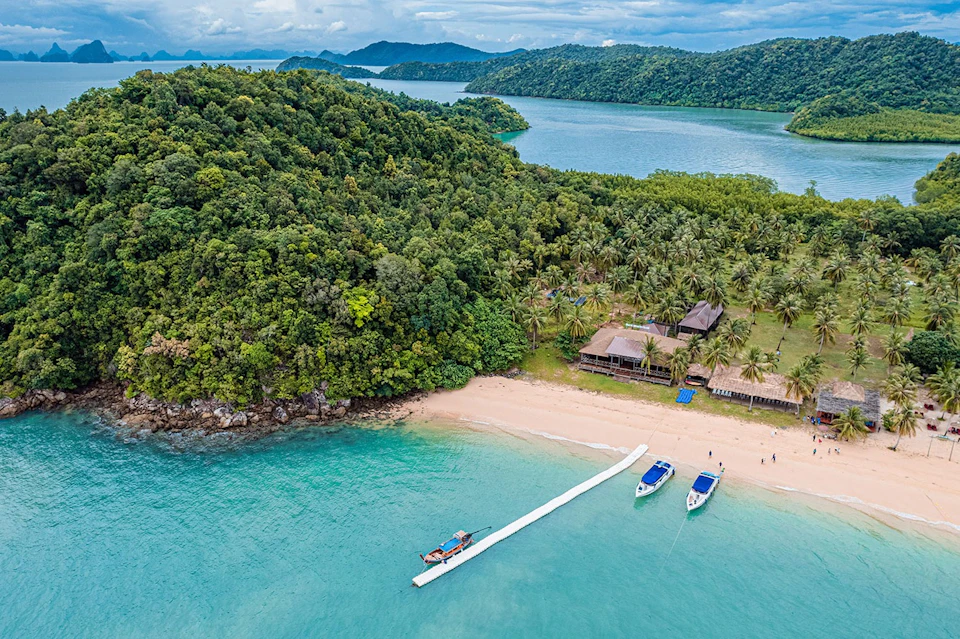 Aerial view of a tropical beach with boats docked at a pier, dense green forest, and buildings near the shoreline.