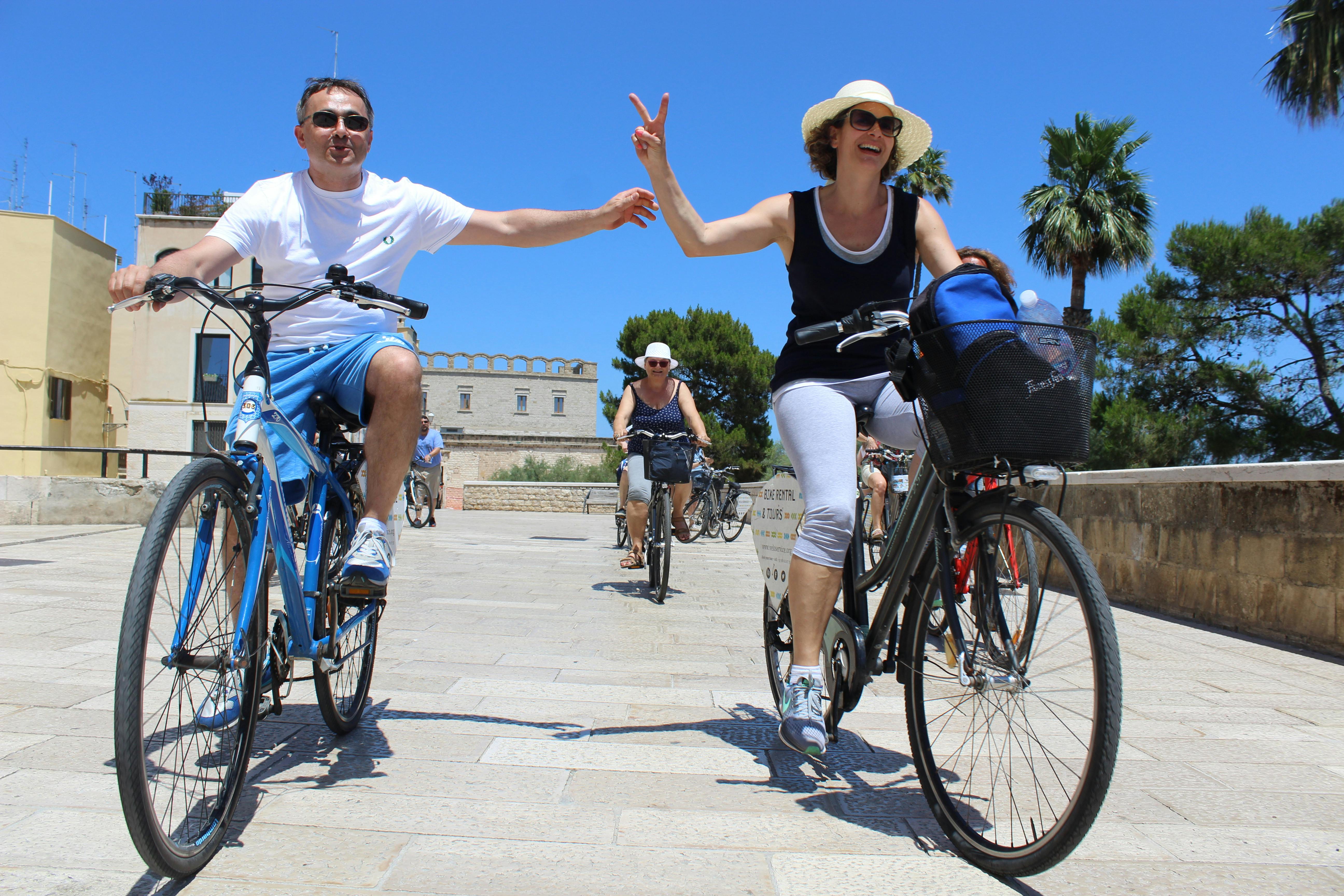 Two people ride bicycles on a sunny day; the man holds the woman's hand, and both smile. Others on bikes and buildings in the background.