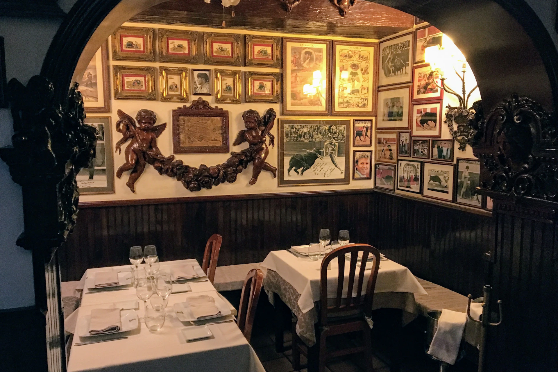 A cozy restaurant corner with framed pictures and artworks on the walls, adorned with cherub sculptures above two set tables.