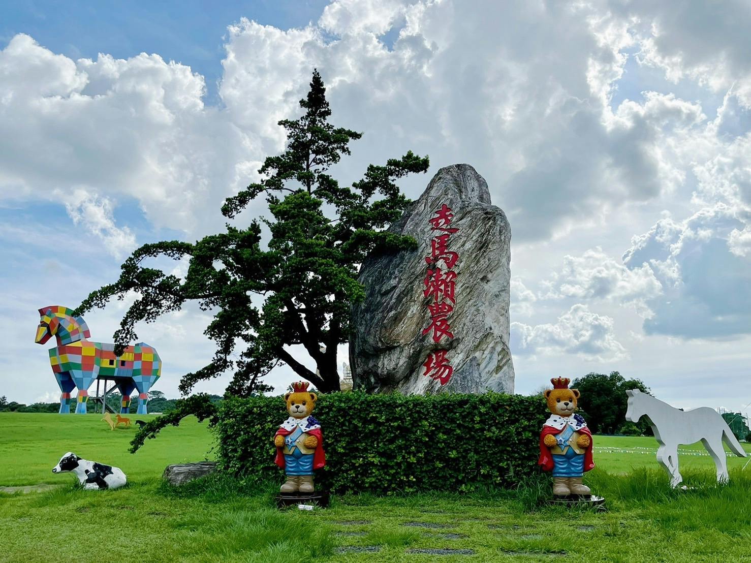 Une scène de parc verdoyant où l'on peut voir une grande statue de cheval aux couleurs vives, une pierre portant une inscription en rouge et deux statues d'ours en tenue royale.