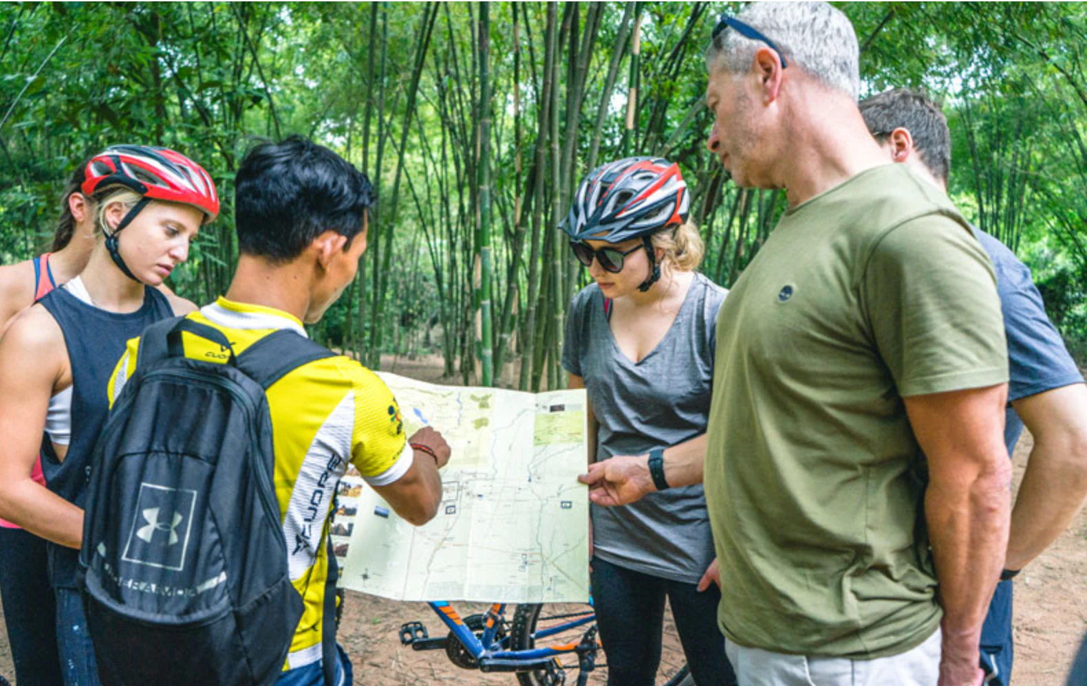 A group of four people in cycling gear examining a map in a bamboo forest, with bikes visible in the background.