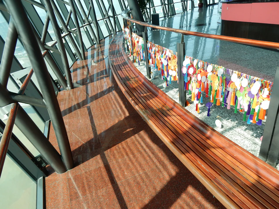 A curved wooden bench beside a railing adorned with colorful hanging decorations inside a modern building with large windows.