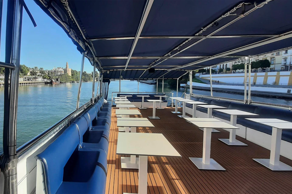Open-air boat with blue canopy, white tables, and seating, cruising on a river with cityscape and trees in the background.