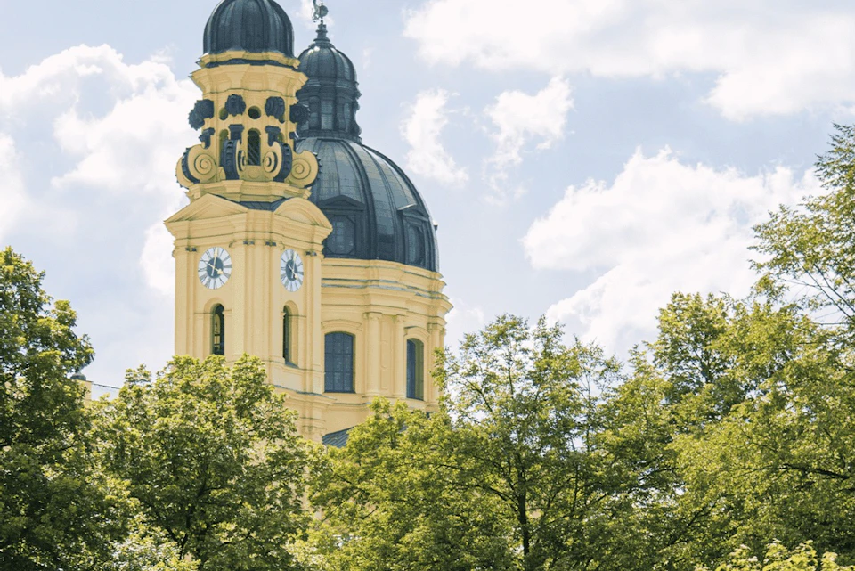 A yellow baroque church tower with a dome rises above green trees under a partly cloudy blue sky.