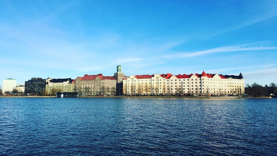 Fila de edificios de colores con tejados rojos a lo largo de un paseo marítimo bajo un cielo azul claro. Agua en primer plano.