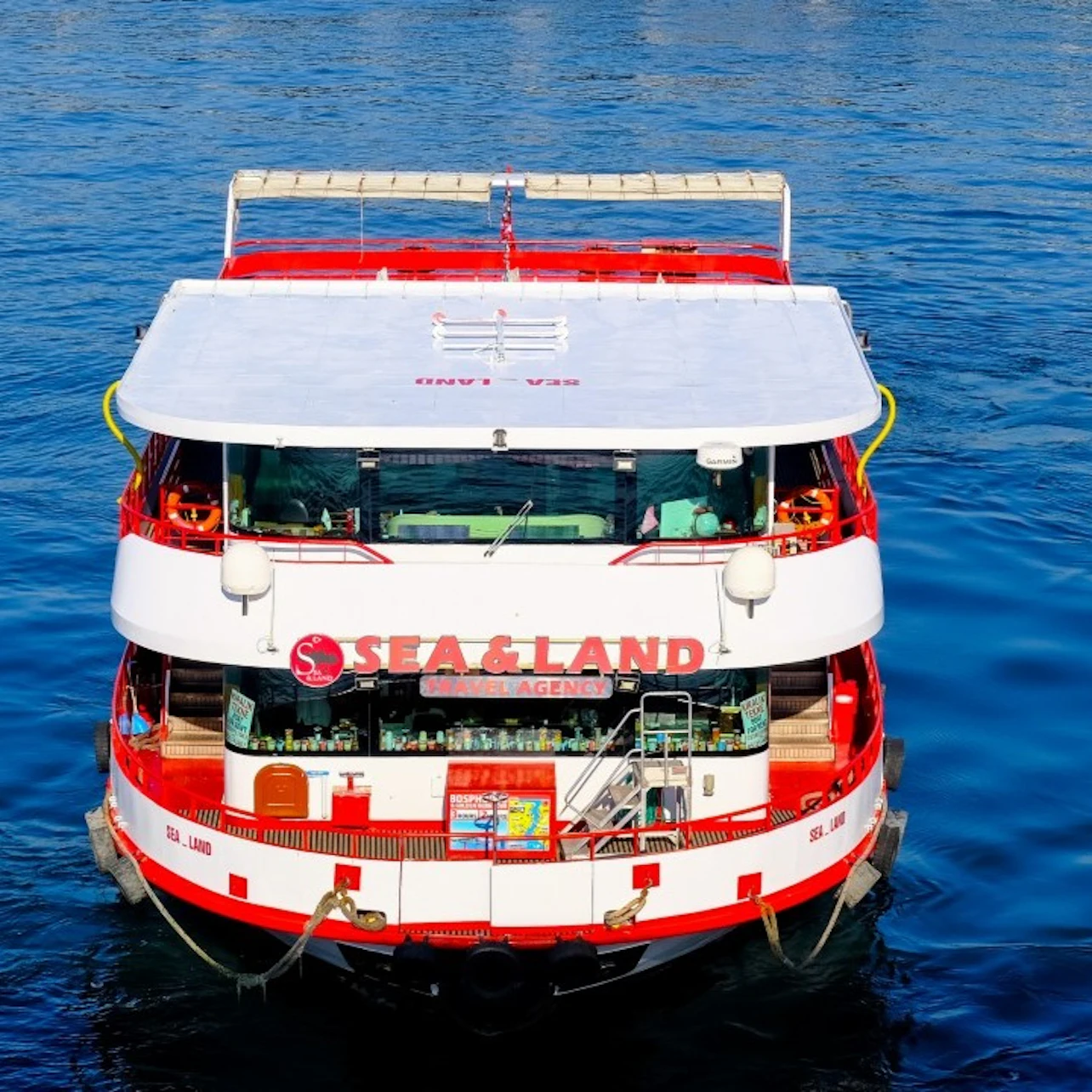A red and white boat named "SEA & LAND" floats on a calm blue water surface, viewed from the rear.
