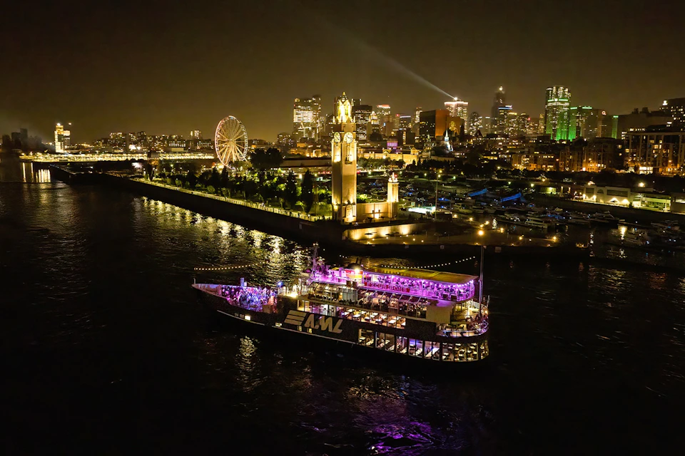 A brightly lit boat on a river at night with a city skyline and an illuminated clock tower in the background.