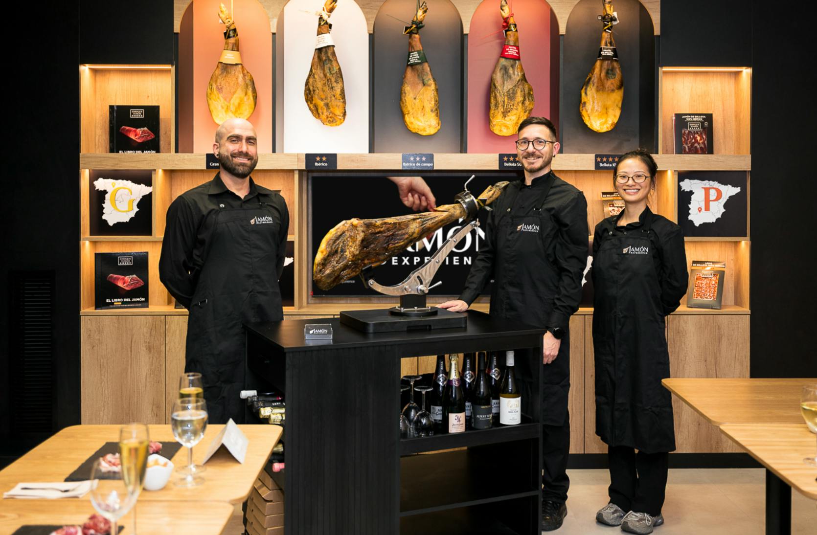 Dois indivíduos em uniformes pretos estão num interior elegante com presuntos curados pendurados, um balcão de exposição e bebidas.