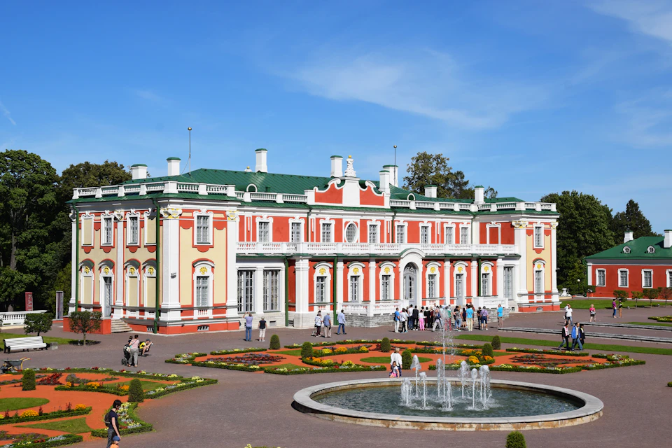 A large Baroque-style building with a green roof, surrounded by gardens and a fountain in front. People are walking and sightseeing.