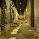 An ancient, dimly-lit stone corridor with weathered brick walls, partially intact arches, and uneven stone flooring.