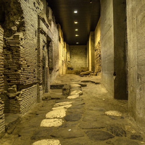 An ancient, dimly-lit stone corridor with weathered brick walls, partially intact arches, and uneven stone flooring.