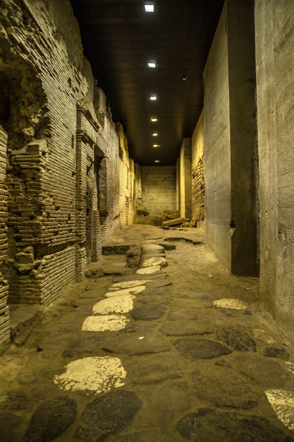 An ancient, dimly-lit stone corridor with weathered brick walls, partially intact arches, and uneven stone flooring.