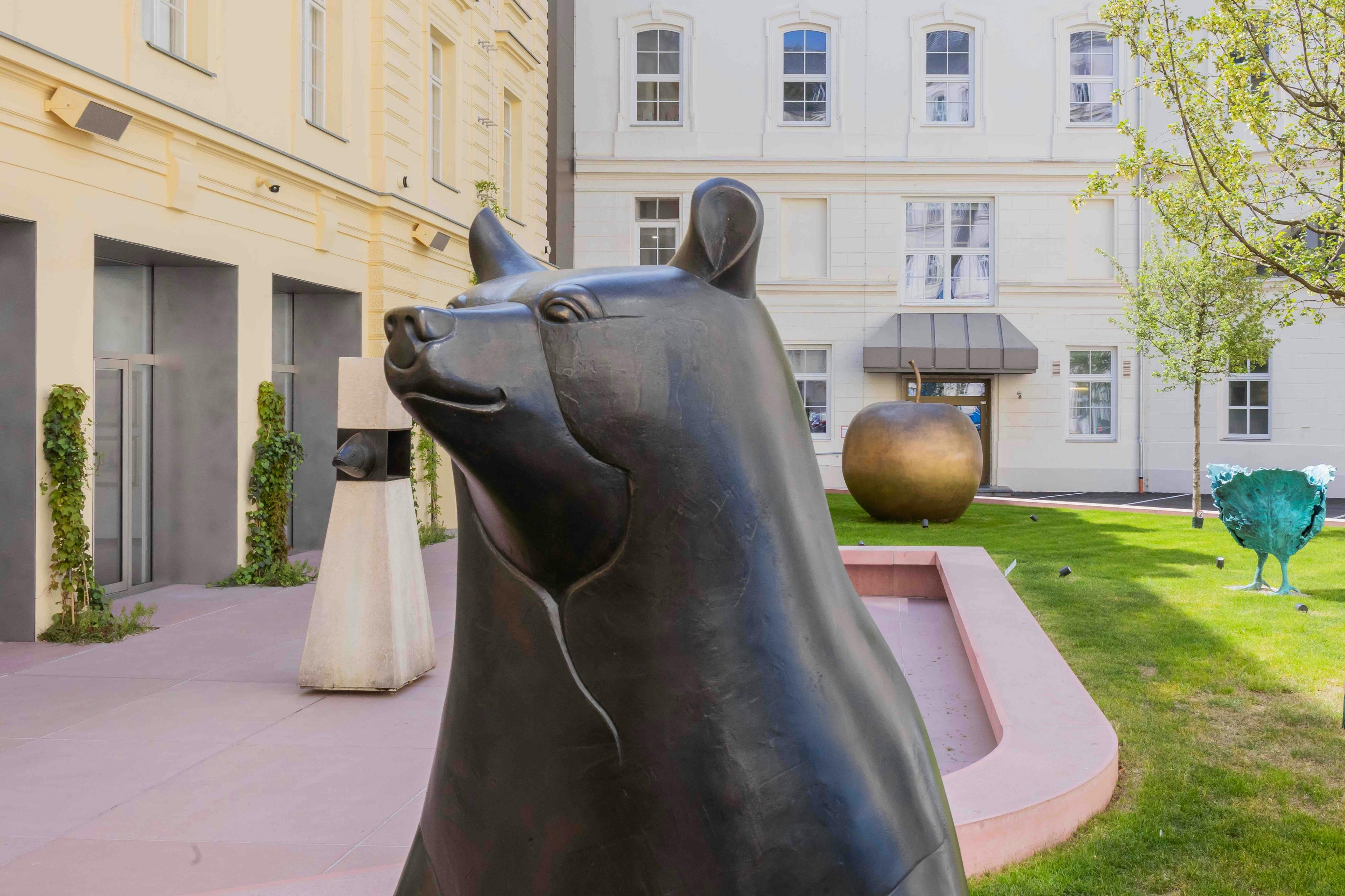 Bronzová medvědí plastika na nádvoří s dalšími abstraktními plastikami a kulisou béžových a bílých budov.