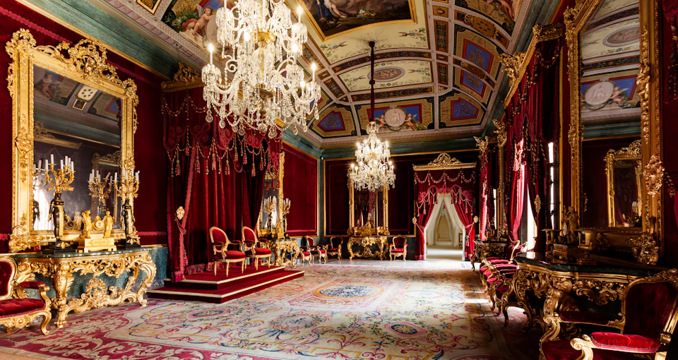 An opulent room with ornate chandeliers, red drapes, gilded chairs, and detailed ceiling paintings, suggesting a throne room.