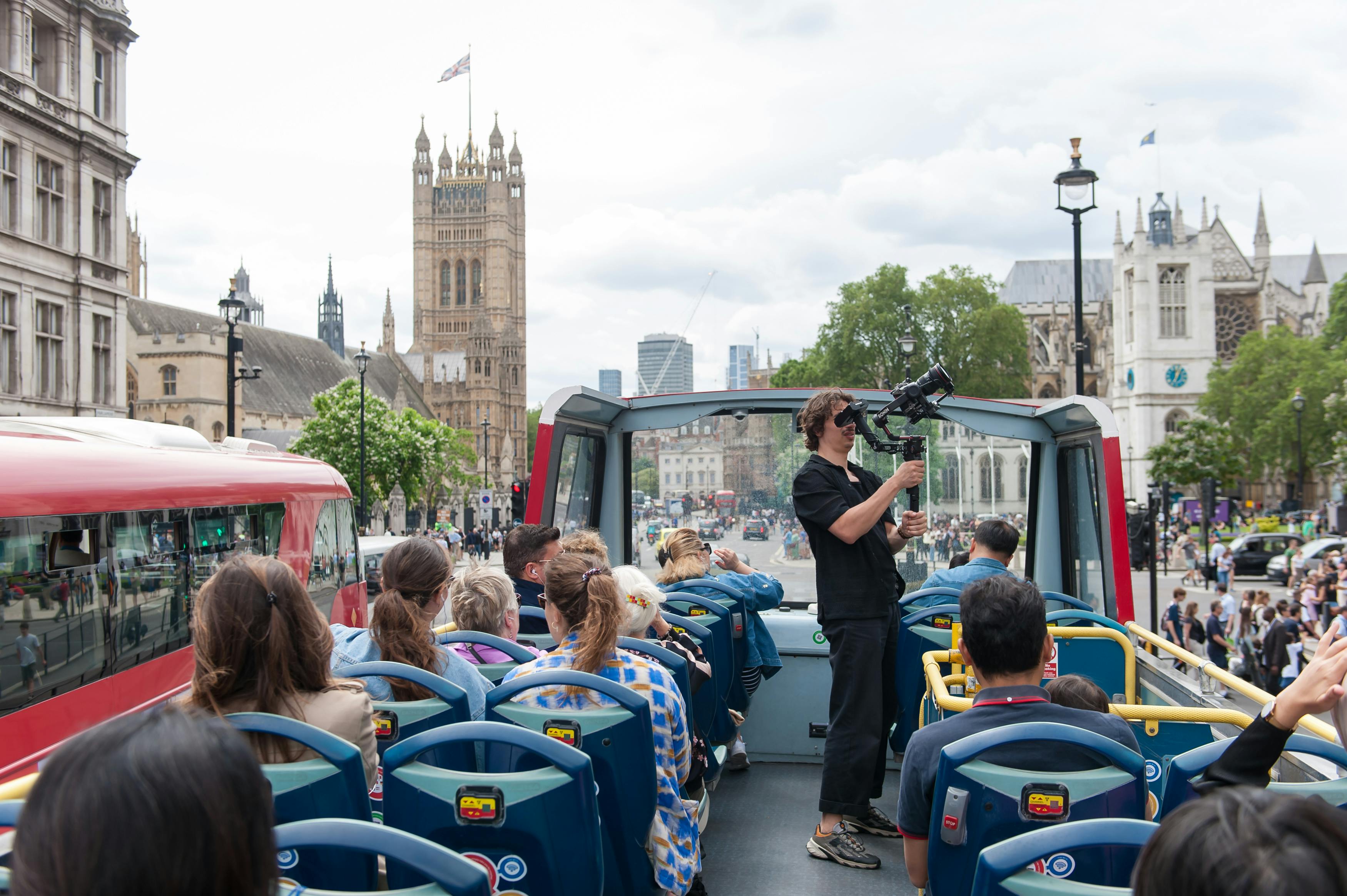 한 사람이 카메라를 이용해 촬영하는 오픈탑 버스 투어에 참여한 관광객들. 거대한 역사적 건물이 배경에 있습니다.