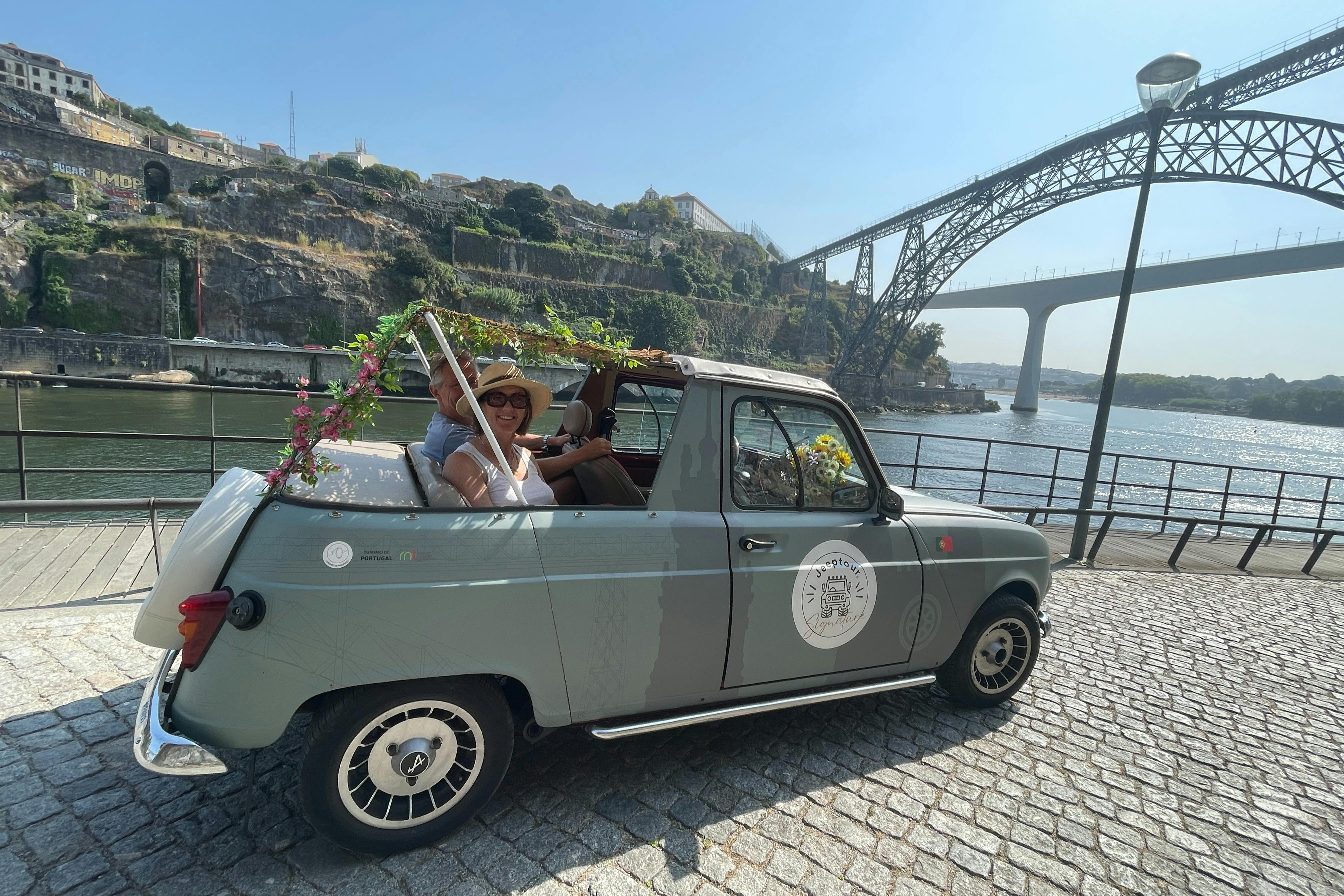 Two people in a vintage, decorated convertible car, parked by a river with a bridge and hillside buildings in the background.