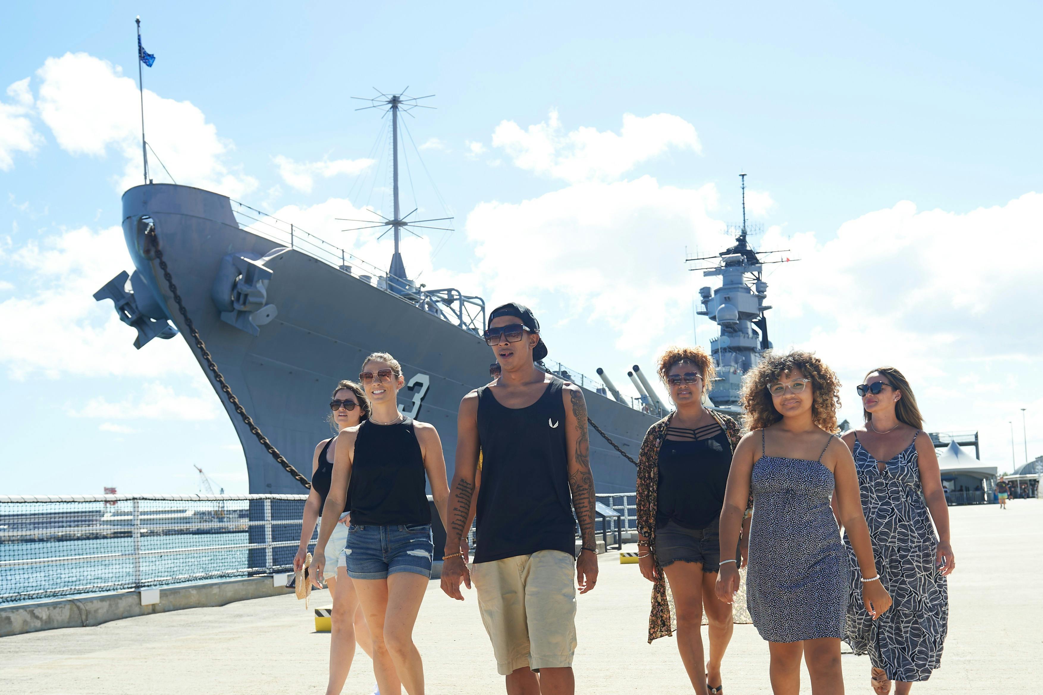 A group of people walking near a large battleship docked at a harbor under a sunny sky.