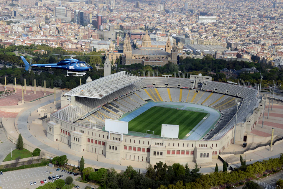 Aerial view of an empty stadium with a blue helicopter flying nearby, historic buildings, and a cityscape in the background.