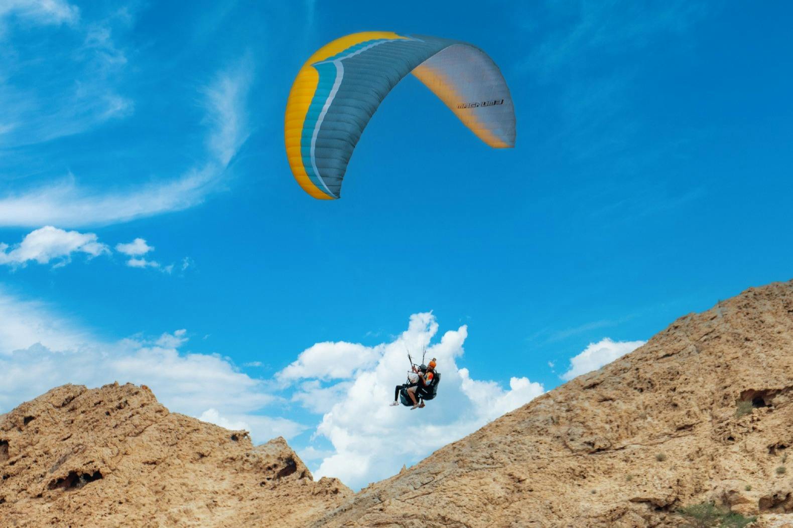 Δύο άνθρωποι κάνουν αλεξίπτωτο πλαγιάς πάνω σε τραχύ, βραχώδες έδαφος με φωτεινό γαλάζιο ουρανό και σύννεφα στο παρασκήνιο.