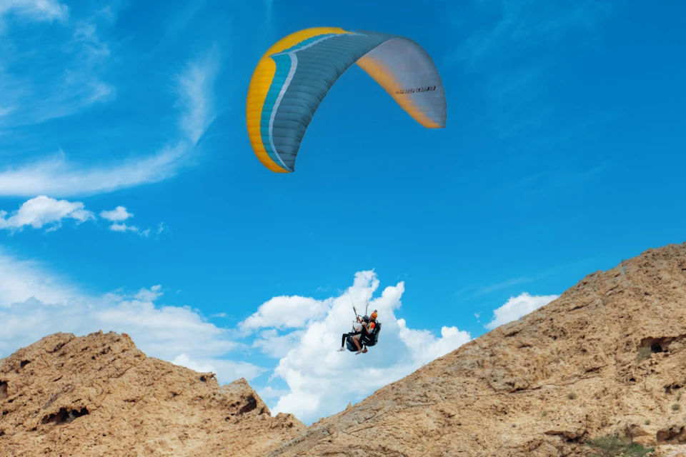 Two people paragliding over rugged, rocky terrain with a bright blue sky and clouds in the background.