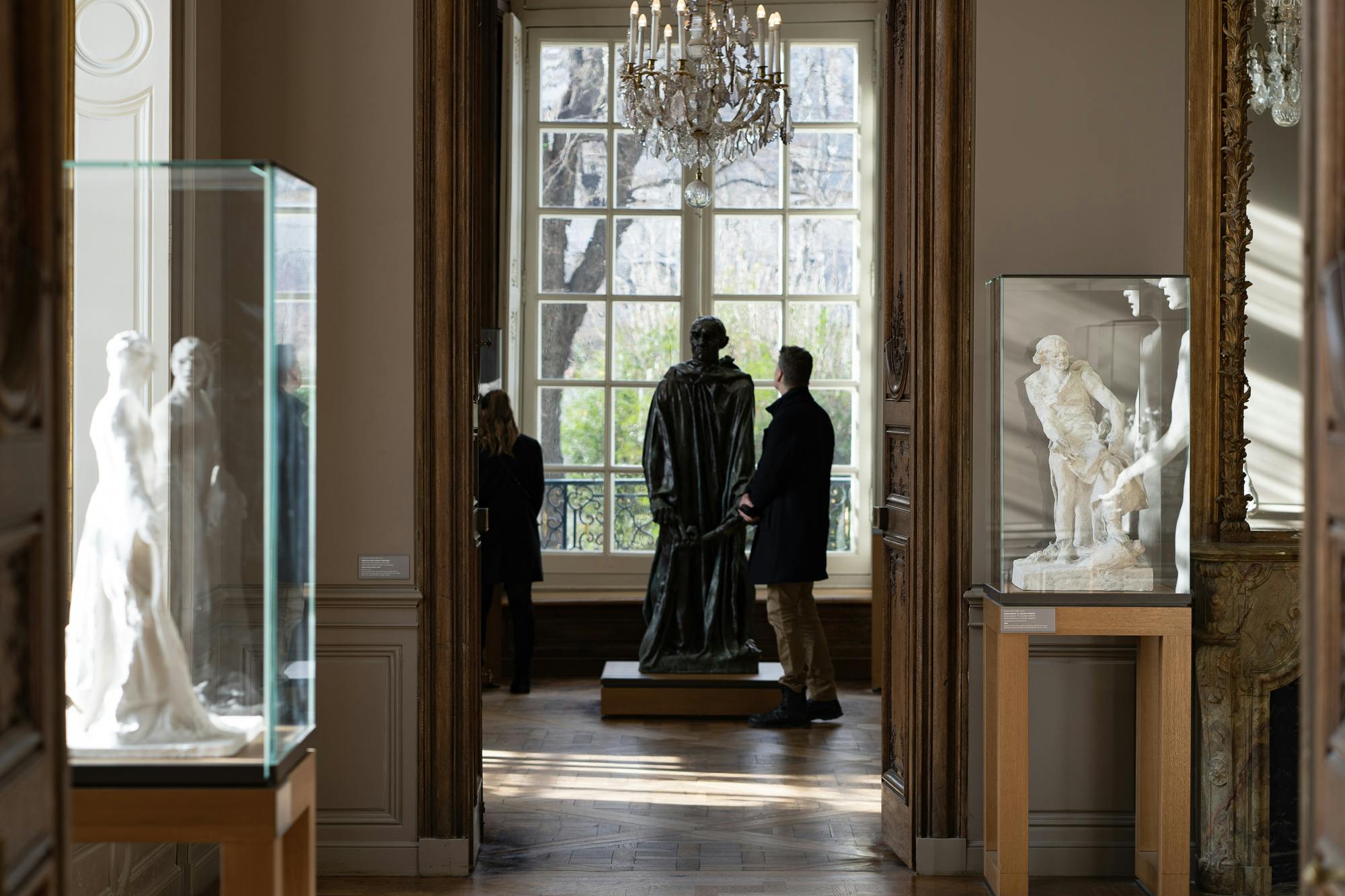커다란 창문, 샹들리에, 유리 케이스에 조각품이 전시된 미술관의 동상 근처에 두 사람이 서 있습니다.
