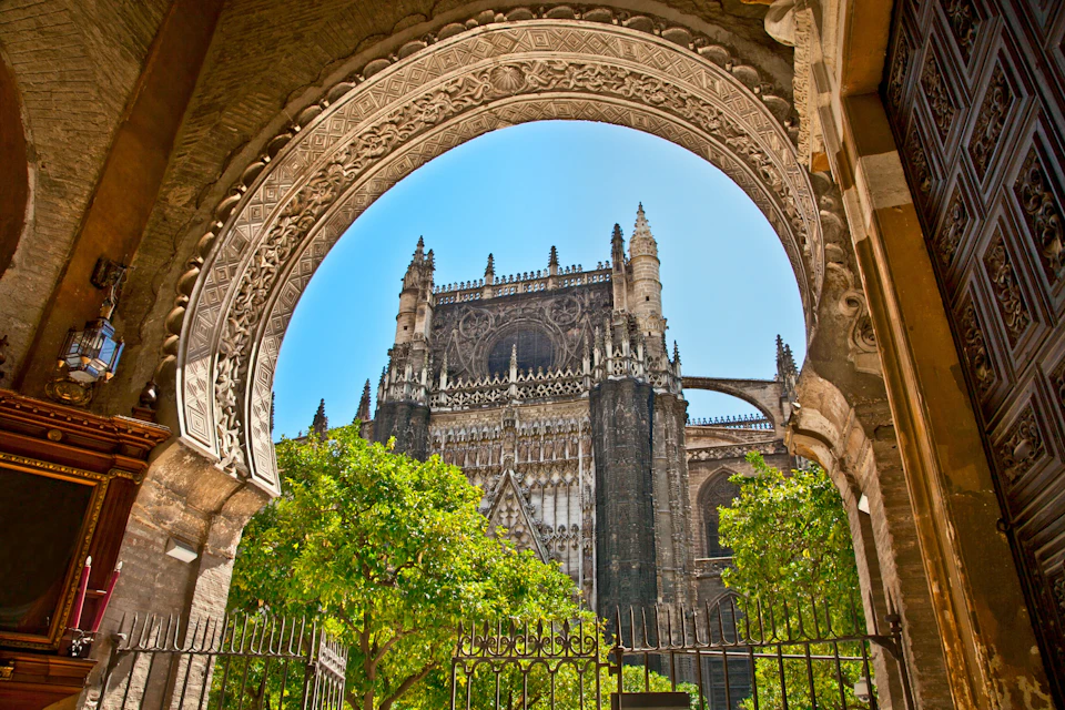 Gothic cathedral framed by an intricately carved archway, with green trees and a metal fence in the foreground.