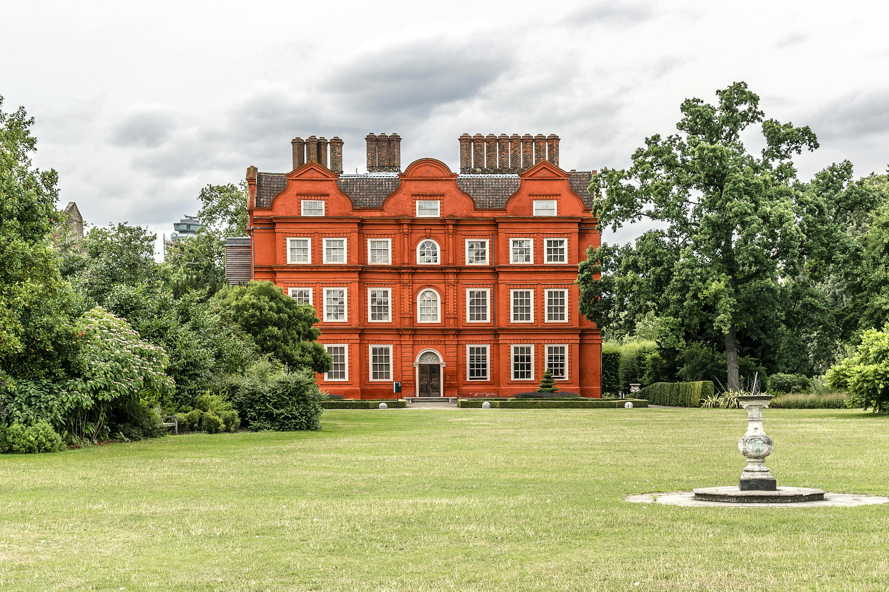 一栋大型红砖建筑，多扇窗户矗立在多云的天空下的绿色草坪和树木之间。