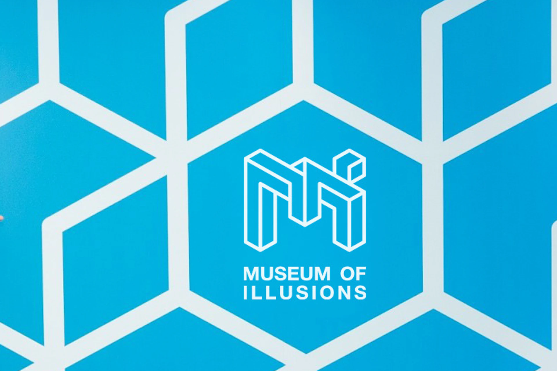 Two people pose playfully against a blue patterned wall at the Museum of Illusions, with part of another person entering the frame.