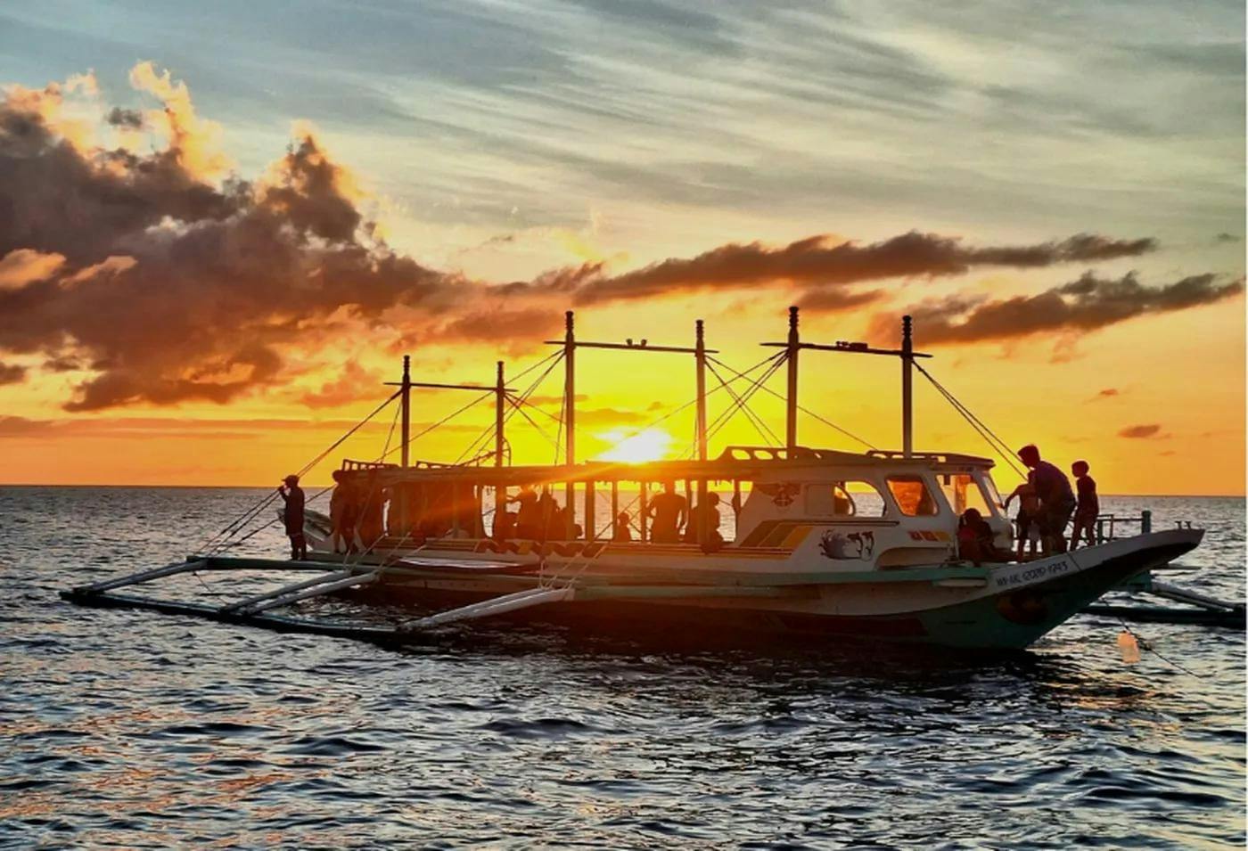 A boat with people on board floating on the water during sunset, with orange and pink clouds in the sky.