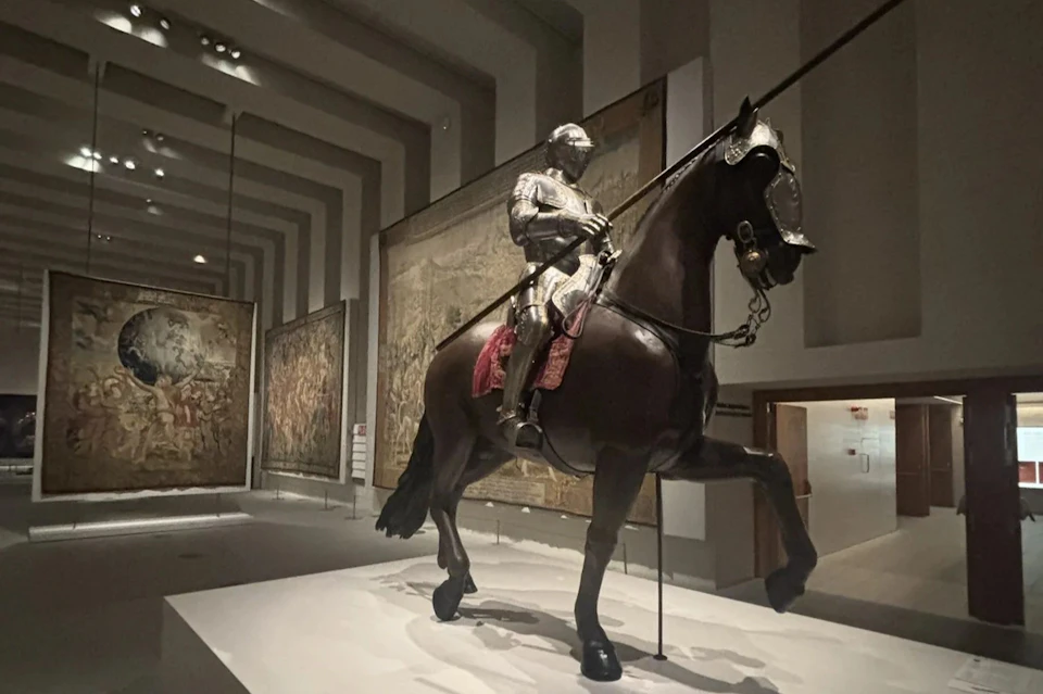 A museum exhibit featuring a knight in full armor on a horse, with tapestries displayed on the walls in the background.