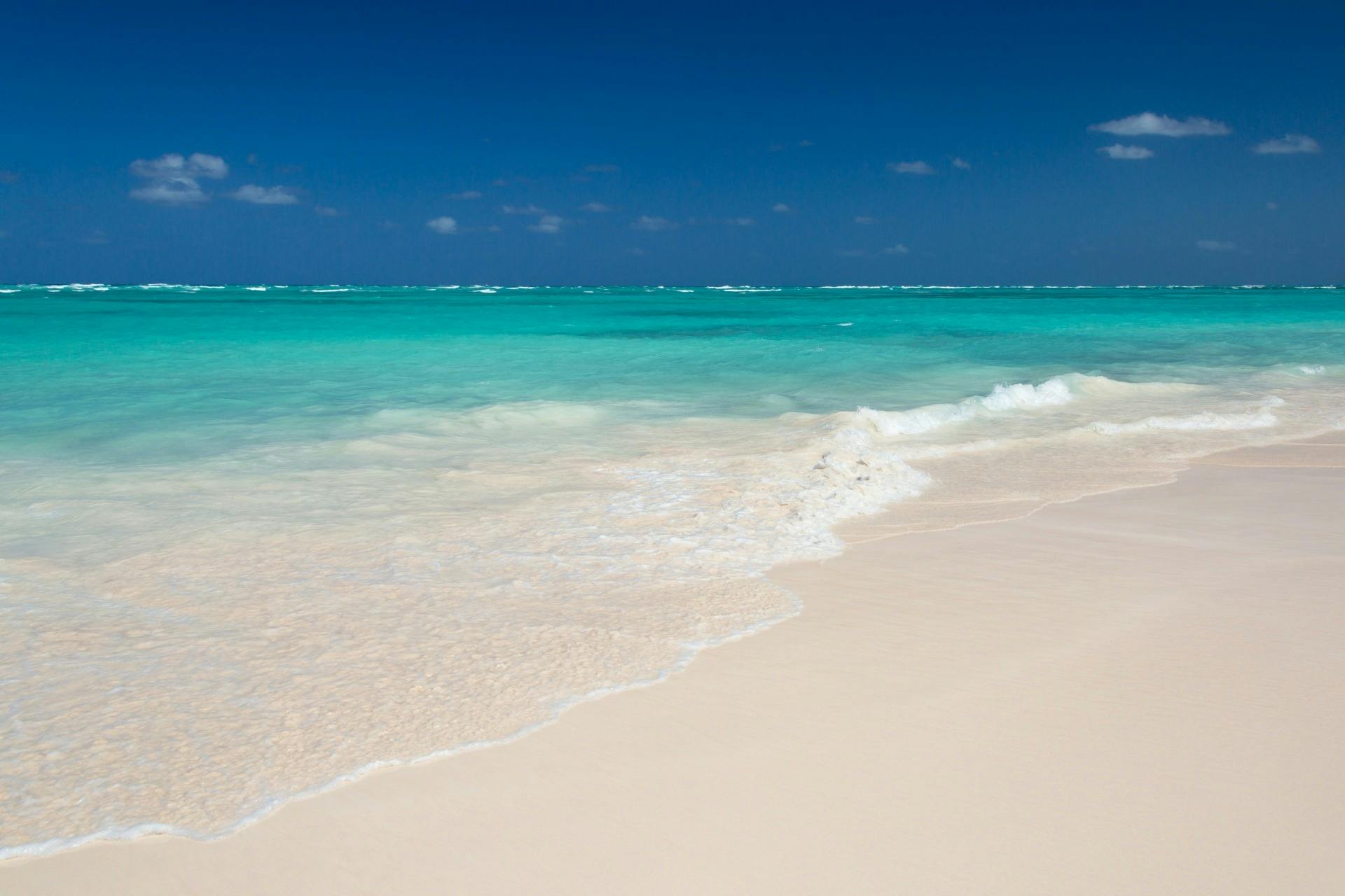 Clear turquoise ocean waves gently lap onto a pristine, sandy beach under a deep blue sky with a few scattered clouds.
