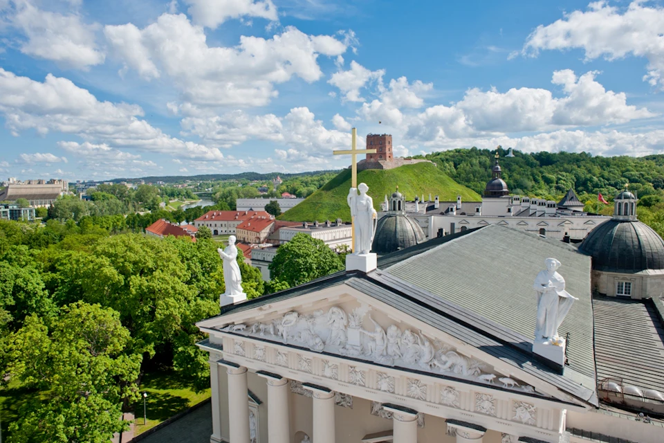 Rooftop view of Vilnius Cathedral with statues and a cross, lush greenery, and Gediminas Tower on a hill under a partly cloudy sky.