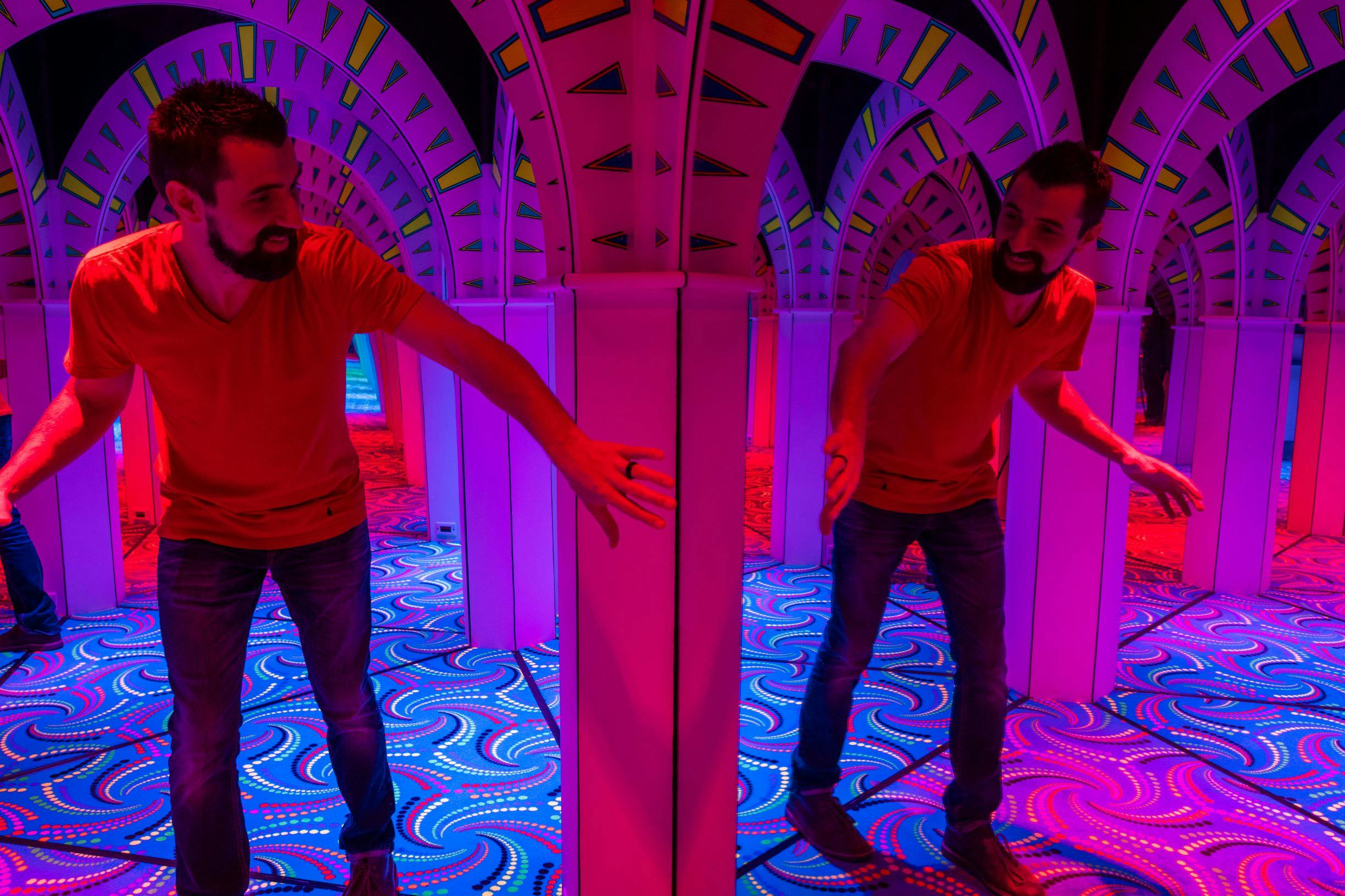 A man in a red shirt navigates a colorful, neon-lit mirror maze, creating multiple reflections.