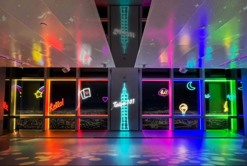 A brightly lit hallway with neon signs, including "Hello!" and "Taipei 101," and cityscape views through large windows.