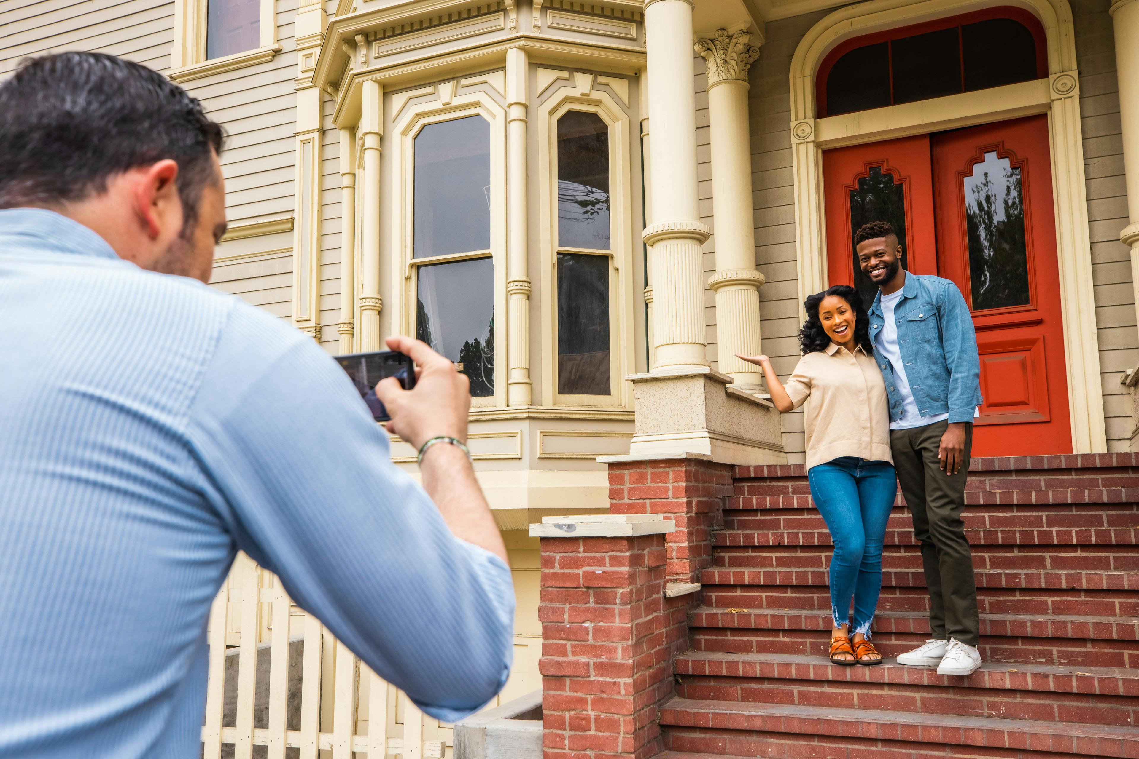 A couple poses for a photo on the steps of a house with red double doors and ornate columns while another person takes their picture.