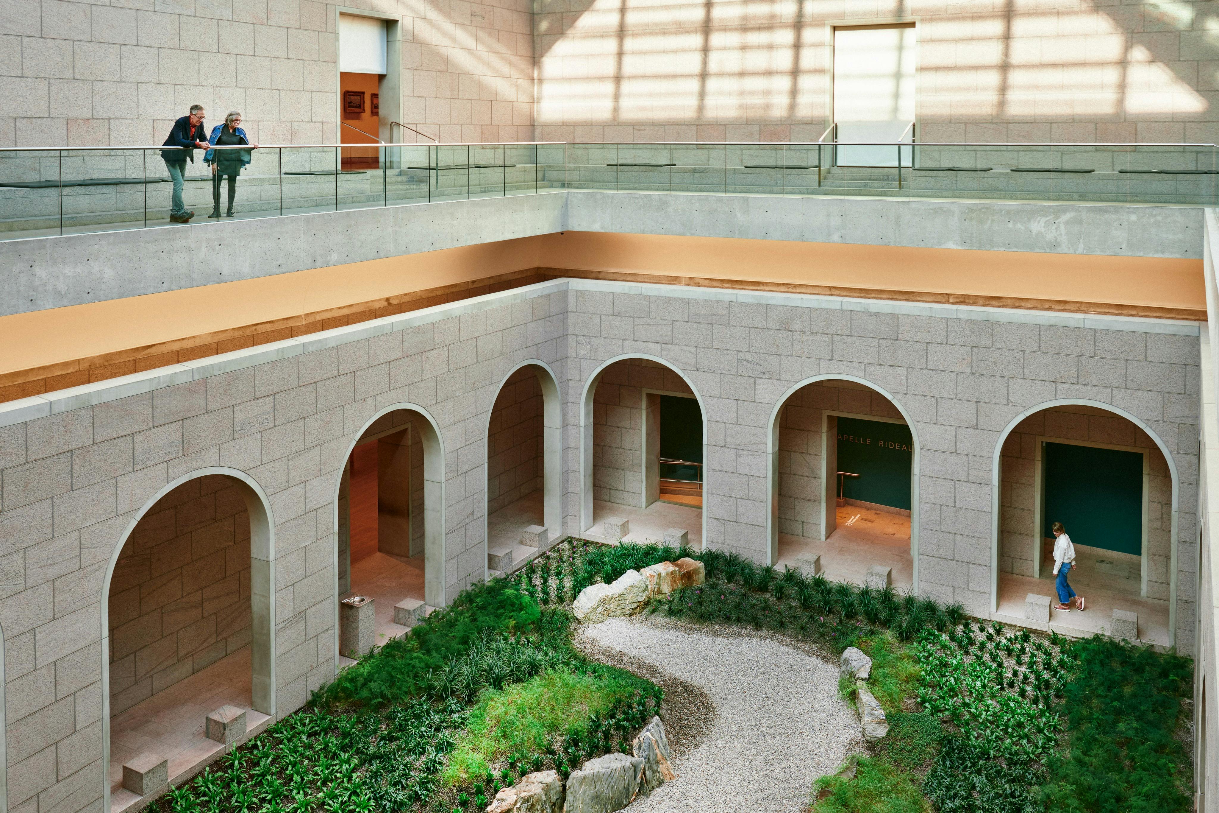Indoor courtyard with arches, a gravel path, greenery, and rocks. A person looks down from the upper level with glass railings.