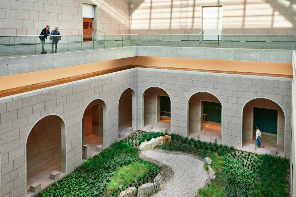 Indoor courtyard with arches, a gravel path, greenery, and rocks. A person looks down from the upper level with glass railings.